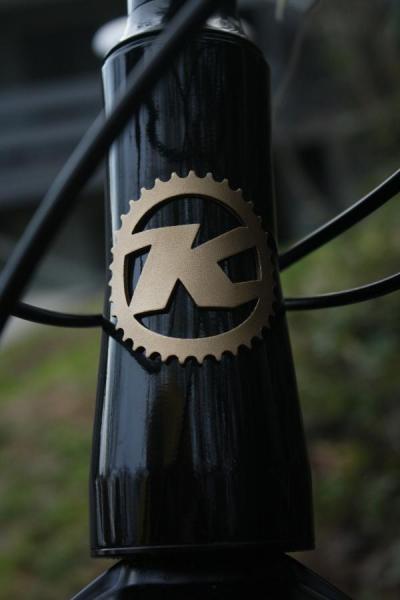 Kona Tanuki: Close-up of a bicycle's head tube featuring a black finish and a bronze logo shaped like the letter "K" within a circular gear design. The background is blurred, emphasizing the logo.
