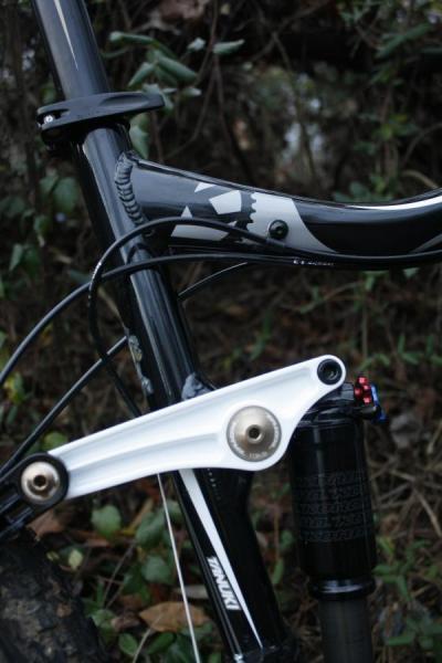 Kona Tanuki: Close-up view of a mountain bike's frame, showcasing the black and white design of the handlebars and the suspension linkage. The image highlights the bike's mechanical components amidst a natural, leafy background.
