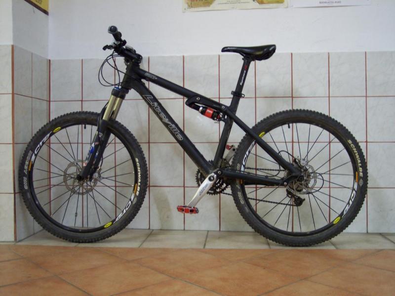 Liteville 301: A black mountain bike leaning against a tiled wall. The bike features thick tires, a front suspension fork, and disc brakes. The setting includes a tiled floor with a subtle pattern and a wall with a map in the background.