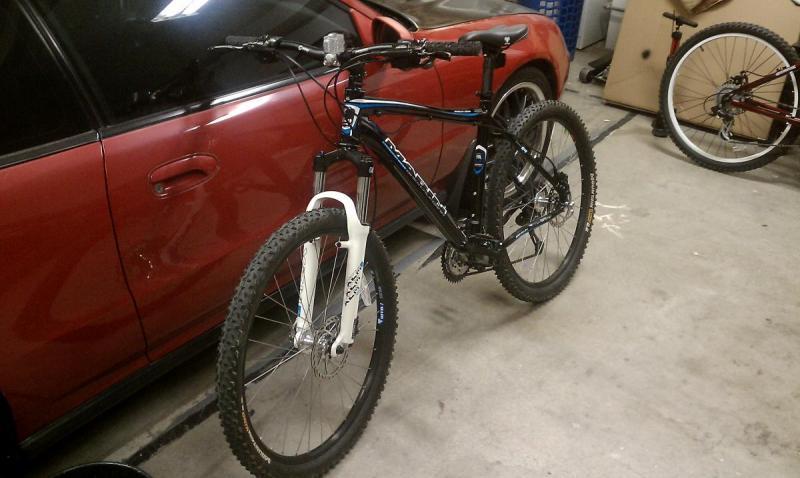 Marin Bobcat Trail: A mountain bike with a black frame and white front fork stands in a garage next to a red car. Other bicycles are visible in the background, and various items are scattered around the floor. The lighting is dim, typical of an indoor garage setting.