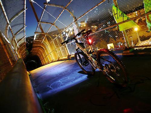 A bicycle parked on a lit section of a fenced bridge at dusk, with city lights visible in the background. The scene features a glowing front light on the bike and colorful decorations along the bridge.