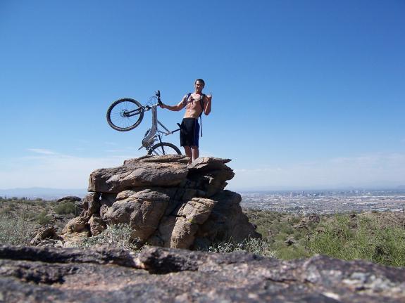 Mongoose Blackcomb: A person stands triumphantly on a rocky outcrop, holding a mountain bike above their head with one arm raised in a celebratory gesture. The backdrop features a clear blue sky and a sprawling view of a valley, suggesting a vibrant outdoor setting. The individual is shirtless and appears energized, embodying the spirit of adventure and achievement.