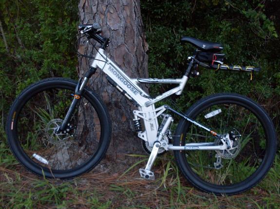 Mongoose Blackcomb: A white mountain bike leaning against a tree, featuring a dual suspension system, wide tires, and a comfortable seat. The bike has various components visible, including gears, pedals, and a small pouch attached to the seat. Surrounding vegetation includes grass and nearby trees.