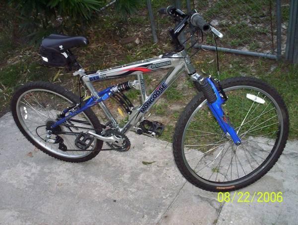 A silver and blue Mongoose mountain bike is positioned on a concrete surface. The bike features front suspension, black handlebars, and a small black saddlebag attached to the rear. The scene includes a grassy area in the background, indicating an outdoor setting. The date displayed in the bottom corner is August 22, 2006.