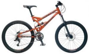 Mongoose Teocali Comp: A mountain bike with an orange frame, featuring wide tires, a suspension system, and a gear mechanism, displayed against a plain background.