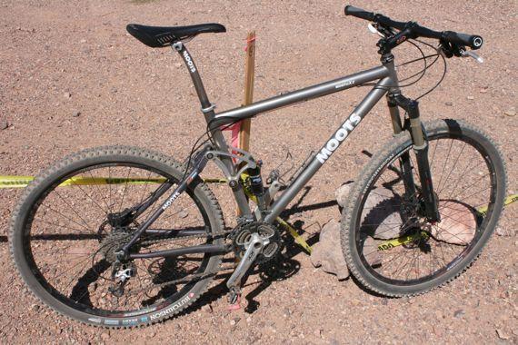 Moots Mooto XZ: A Moots mountain bike resting on gravel terrain, showcasing its sleek gray frame, dirt-covered tires, and visible components like the gears and brake system. The bike is positioned near boundary markers on the ground.