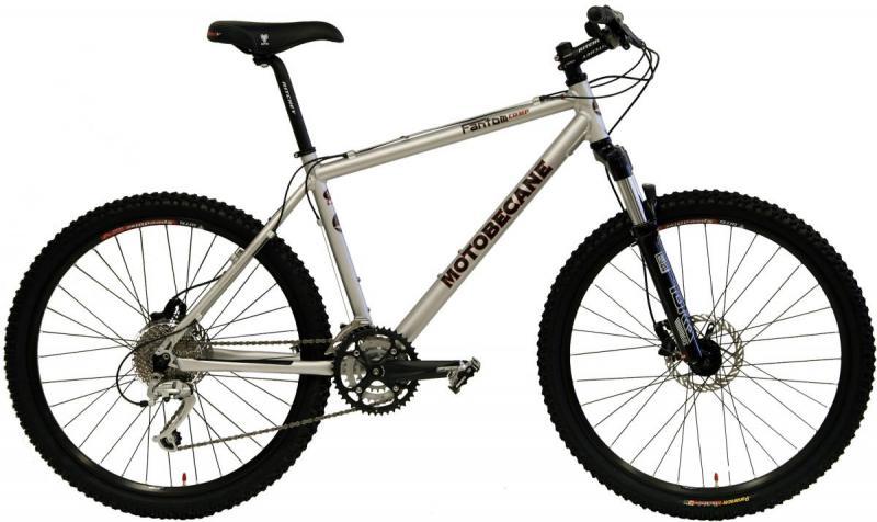Motobecane Fantom Comp: A silver Motobecane mountain bike with a sturdy frame, front suspension, and thick black tires, positioned against a plain white background. The bike features a multi-speed gear system, disc brakes, and a comfortable saddle designed for off-road riding.