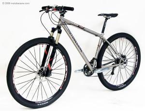 Motobecane Fly Team Ti 29er: A silver and black mountain bike with thick tires, front suspension, and a modern frame design, displayed against a plain white background. The bike features a sleek, lightweight build suitable for off-road terrain.