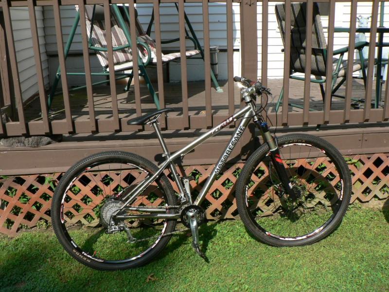 Motobecane Fly Team Ti 29er: A mountain bike with a silver frame is leaning against a wooden deck railing. The bike has high-performance tires and is positioned on a grassy area, with a glimpse of a patio in the background featuring a swing and chairs.