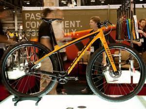 Niner Air 9 Carbon: A black and orange mountain bike displayed on a stand at an exhibition, with blurred figures of people in the background and various colorful bike accessories visible on the back wall.