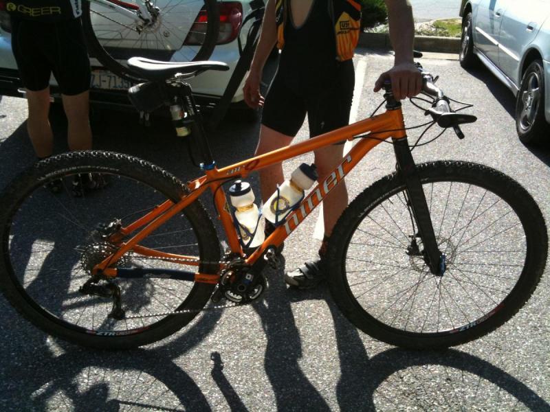 Niner Air 9: Mountain bike with an orange frame parked on a paved surface, featuring two water bottle holders. A person in cycling attire stands next to the bike, with part of their leg and arm visible. In the background, a car is partially visible. The scene is sunny and captures a moment before a ride.