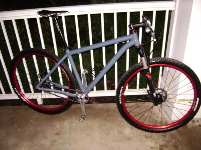 Niner Air 9: A gray mountain bike with red accents, featuring thick tires, front suspension, and a minimalist frame, parked on a concrete surface next to a white railing. The background shows blurred greenery.