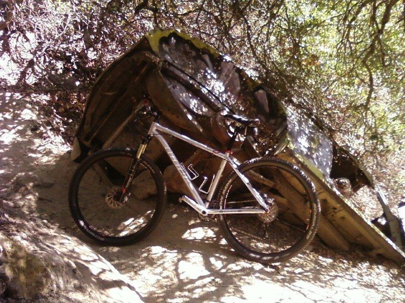 Niner Air 9: A mountain bike resting on a dirt path next to an abandoned, overturned vehicle, surrounded by trees and foliage. The bike has a silver frame and is positioned to the right of the image, while the vehicle, showing signs of rust and age, leans against the natural landscape.