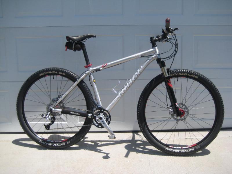 Niner Air 9: A silver and black mountain bike leaning against a garage door. The bike features thick, knobby tires and a lightweight frame. The handlebars have grip tape and a brake lever, while the bike has a single front suspension fork. The scene is well-lit and shows the bike in a clear, side view.