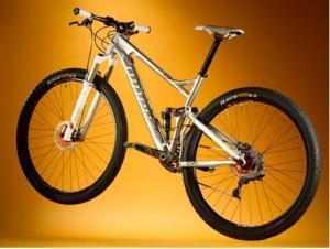 Niner Jet 9: Mountain bike displayed against a gradient gold background. The bike features a sleek silver frame, thick black tires, and prominent suspension components. The image highlights the bike's design and attributes, showcasing its gearing and tire tread.