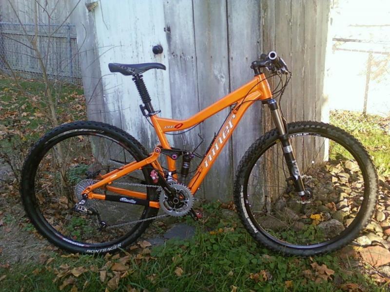 Niner RIP 9: A bright orange mountain bike is positioned against a wooden wall, with its wheels on a patch of grass and fallen leaves around it. The bike features a sturdy frame, front suspension fork, and thick tires designed for off-road trails. The background includes a fence and additional greenery, suggesting an outdoor setting.