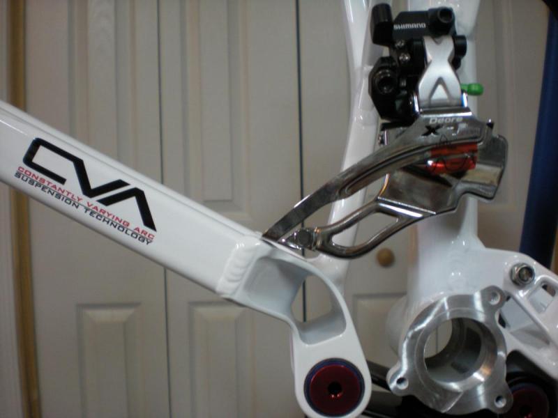 Niner W.F.O. 9: Close-up of a white bicycle frame featuring a Constantly Varying Arc (CVA) suspension design. The image shows the derailleur and chain guide, highlighting the intricate components of the bike's gearing system. The background consists of a neutral setting with light-colored walls, emphasizing the bike parts.