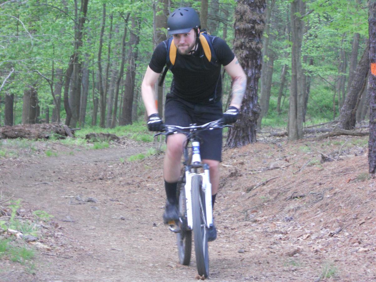 A mountain biker wearing a helmet and black attire rides on a dirt trail surrounded by greenery and trees. The cyclist is in motion, demonstrating an active outdoor lifestyle. North Park mountain bike trail.