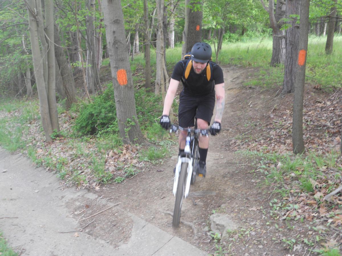 A person riding a mountain bike along a dirt trail through a wooded area, surrounded by trees with orange markings. The cyclist is wearing a helmet and gloves, and the path is a mixture of dirt and gravel. North Park mountain bike trail.
