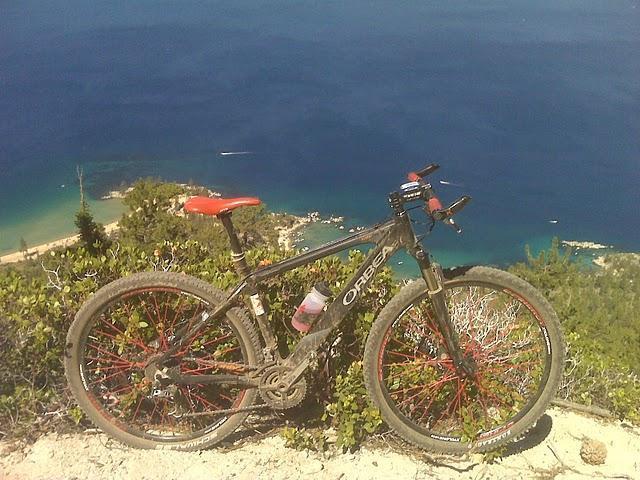 Orbea Alma 29 World Cup: A mountain bike resting on rocky terrain with a scenic view of a blue ocean and coastline in the background. The bike features a black frame with red accents and is positioned amidst greenery, highlighting a beautiful outdoor setting.