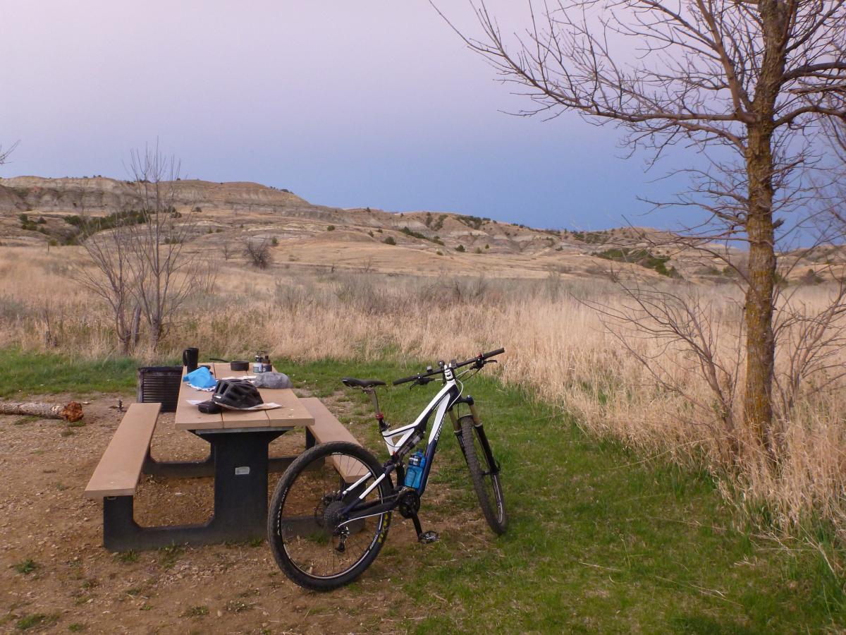 A mountain bike parked next to a picnic table in a grassy area, with a backdrop of rolling hills and a partly cloudy sky. Nearby items on the table include a helmet, a cup, and some personal belongings, suggesting a break during a biking excursion in a natural setting. Maah Daah Hey mountain bike trail.