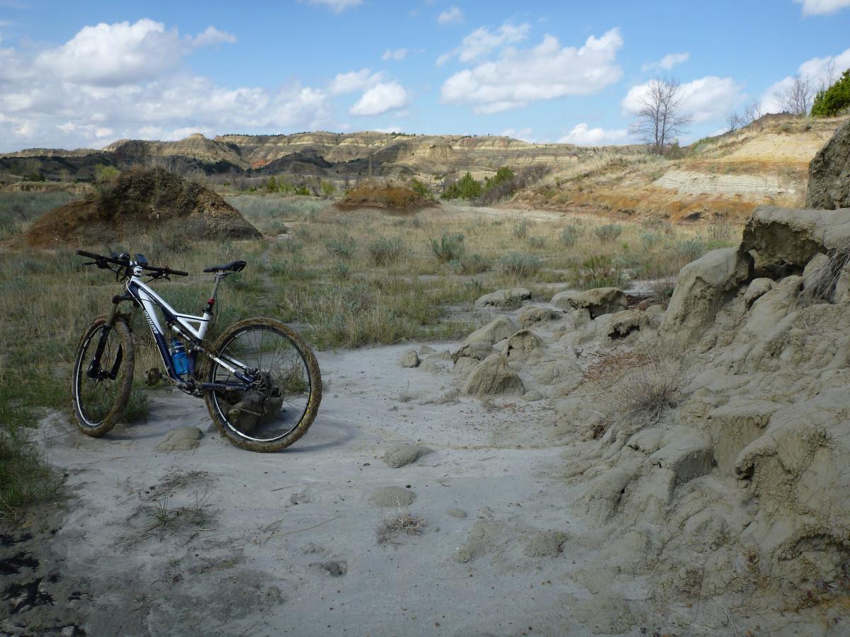A mountain bike resting on sandy terrain surrounded by hilly landscapes with sparse vegetation under a partly cloudy sky. Maah Daah Hey mountain bike trail.