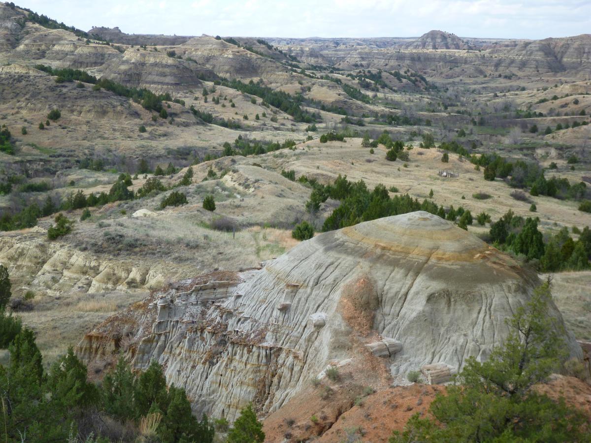A scenic view of rolling hills and layered rock formations in a dry landscape, featuring a prominent conical rock in the foreground. The terrain is mostly barren with patches of green foliage, under a partly cloudy sky. Maah Daah Hey mountain bike trail.