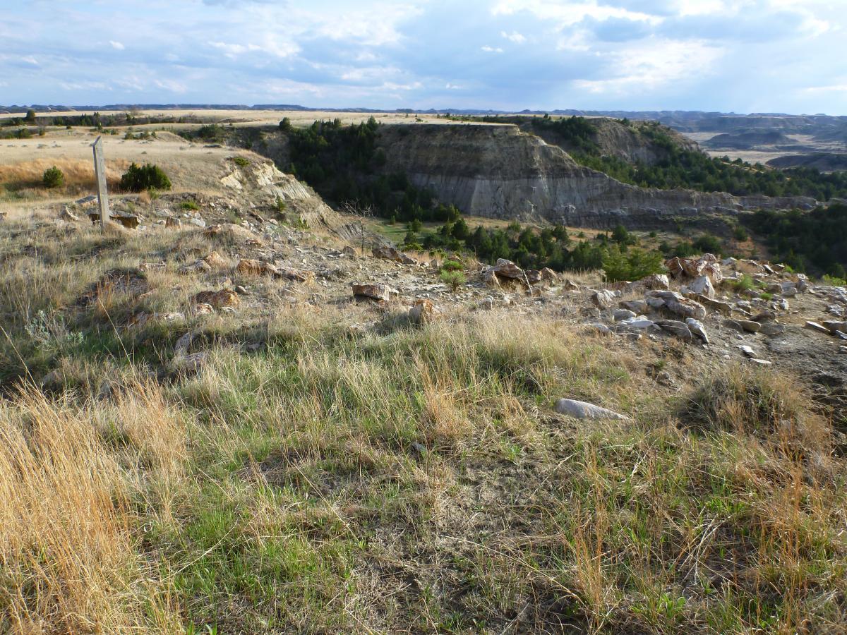 A panoramic view of a rugged landscape featuring grassy hills and rocky terrain, with a deep canyon in the background. The sky is partly cloudy, and a wooden post is visible on the left side of the image. The scenery suggests a remote, natural environment, likely in a rural area. Maah Daah Hey mountain bike trail.