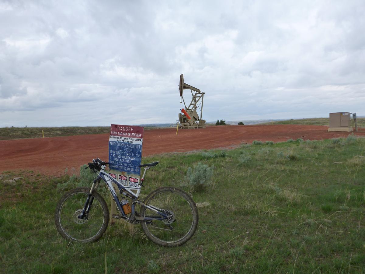 A mountain bike resting on a sign that warns of potential gas hazards, with an oil pump jack visible in the background against a cloudy sky. The terrain is a mix of red dirt and green grass, indicating an industrial area in a rural setting. Maah Daah Hey mountain bike trail.