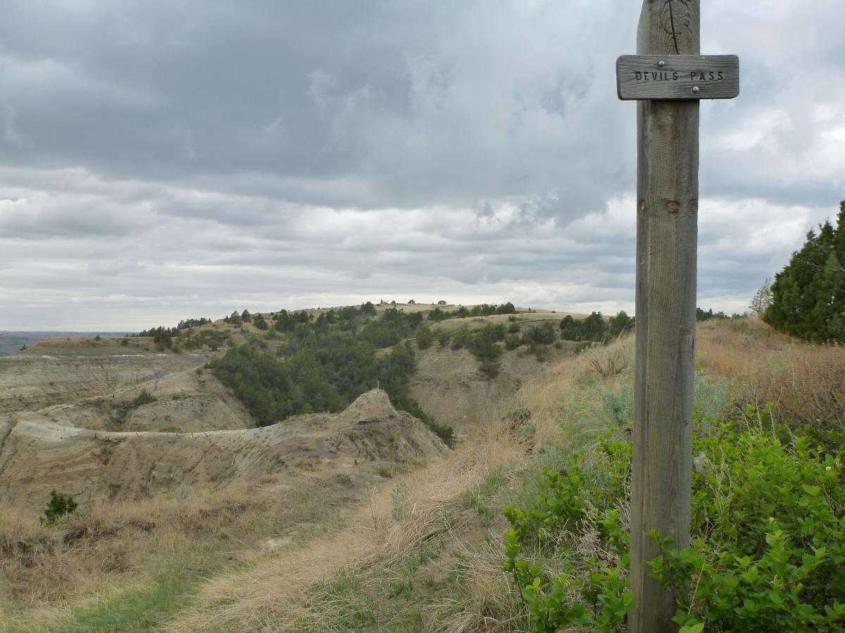 A scenic landscape view of Devil's Pass, featuring a wooden sign with the name "Devil's Pass" at the forefront. The background showcases rolling hills and rugged terrain, with sparse greenery and scattered trees under a cloudy sky. Maah Daah Hey mountain bike trail.