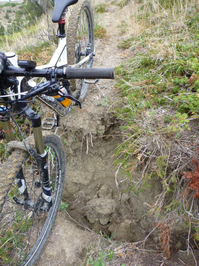 A close-up view of a mountain bike on a rugged trail, with the front wheel positioned near a deep rut in the ground. The surrounding area features sparse vegetation and dirt, indicating a challenging biking path. Maah Daah Hey mountain bike trail.