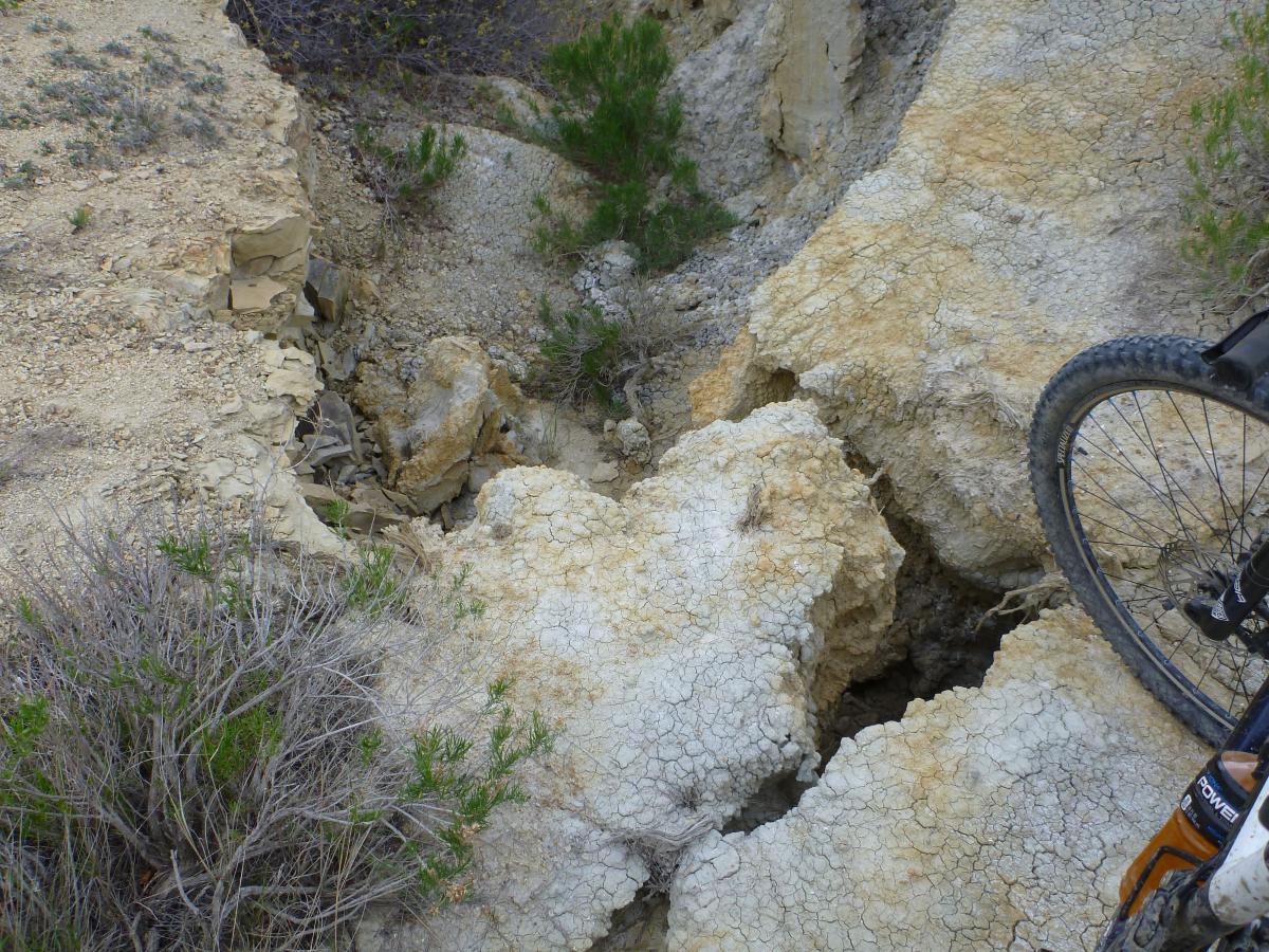 A rocky, dry landscape with cracked earth and scattered vegetation, featuring a steep crevice leading into the ground. A partial view of a bicycle wheel is visible on the right side of the image, indicating an outdoor activity in a rugged terrain. Maah Daah Hey mountain bike trail.