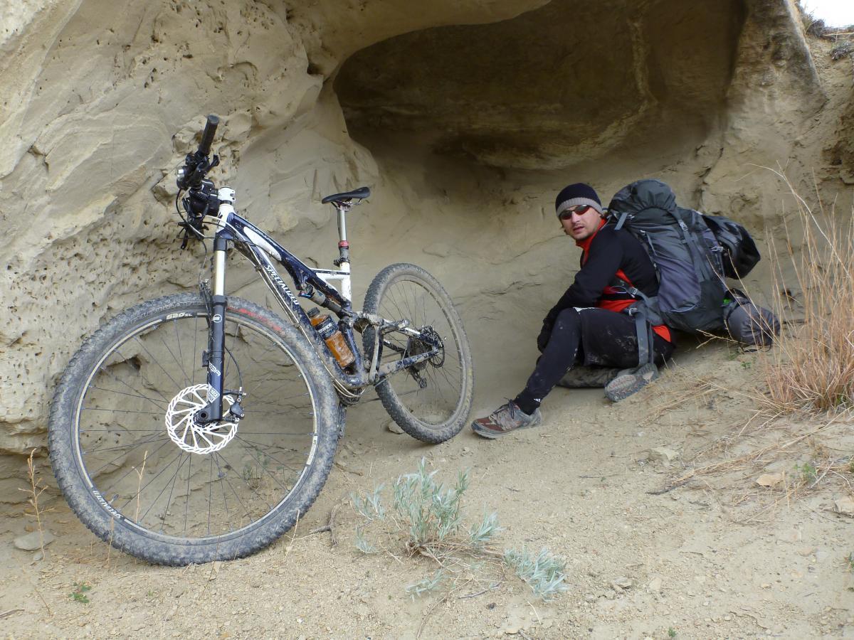 A person resting with a large backpack next to a mountain bike, positioned under a rocky overhang. The individual is wearing a black beanie and sunglasses, dressed in a long-sleeve shirt and pants, with dirt on their clothing. The background features a sandy terrain with sparse vegetation and textured rock formations. Maah Daah Hey mountain bike trail.