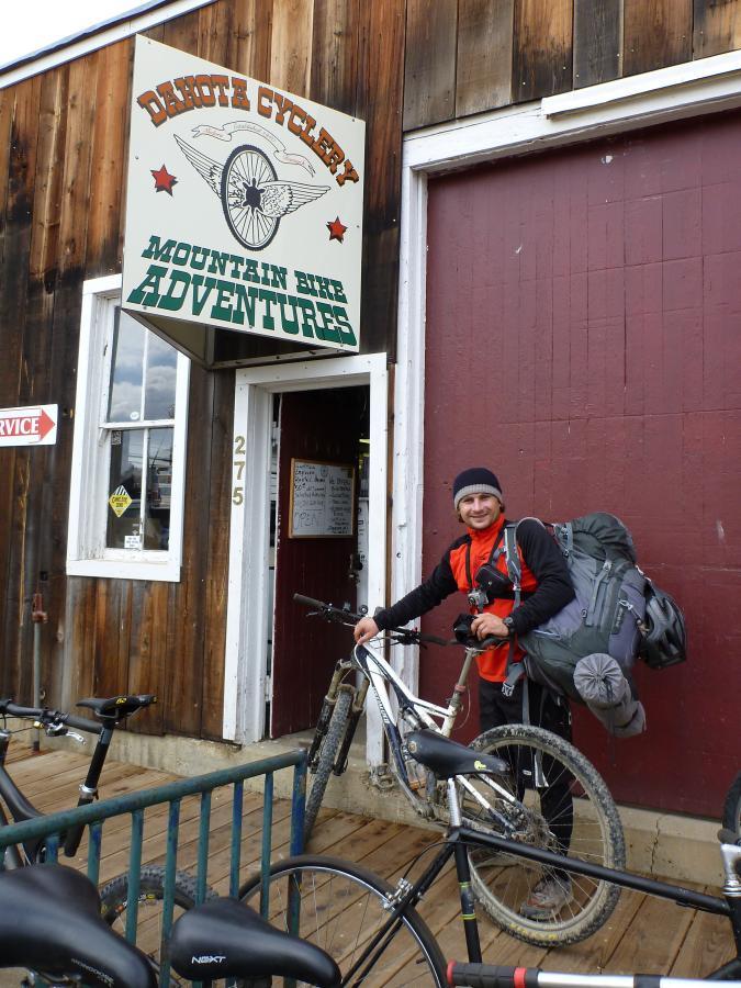 A man with a backpack and camera stands next to a mountain bike in front of Dakota Cyclery, a shop with a wooden facade and a sign promoting mountain bike adventures. The scene includes bikes parked on a wooden deck and a red wall in the background. Maah Daah Hey mountain bike trail.