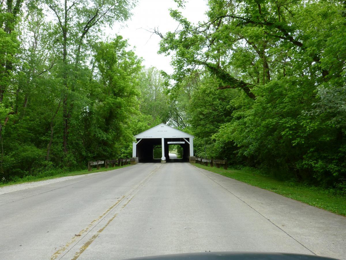A narrow, tree-lined road leading to a white covered bridge, surrounded by lush green foliage. The scene captures a tranquil, scenic view, inviting travelers to pass through the bridge. Brown County Park mountain bike trail.