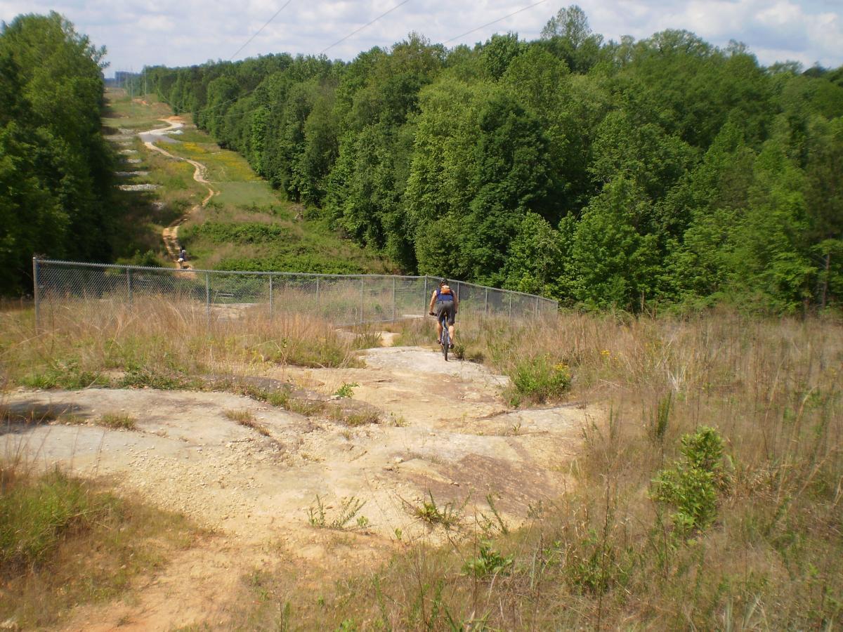 A person riding a mountain bike on a rocky trail surrounded by grass and trees, with a fence in the foreground and a winding path visible in the background. Blue skies with clouds are overhead. Harbins Park mountain bike trail.
