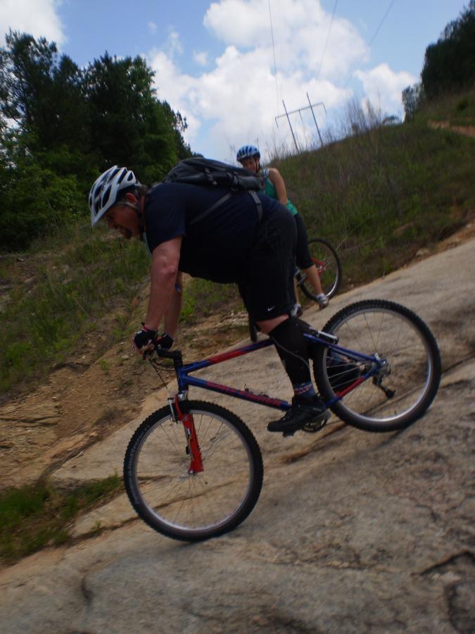 Two mountain bikers riding on a rocky trail surrounded by greenery and under a partly cloudy sky. One rider, wearing a helmet and a backpack, leans forward on a blue and red bike, while the second rider, in a green tank top, follows closely behind. Power lines are visible in the background. Advanced Loop mountain bike trail.