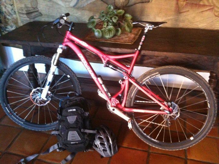 Pivot Mach 429: A red mountain bike is leaning against a wooden table, next to a black backpack and a bicycle helmet. The background features a decorative plant on the table and a patterned wall. The floor is tiled with warm-toned tiles.