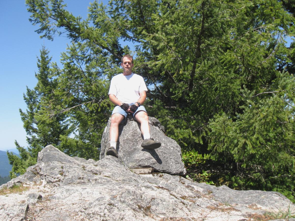 A person sitting on a large rock in a mountainous area, surrounded by lush green trees under a clear blue sky. The individual is wearing a white t-shirt and denim shorts, with their legs casually positioned. Larison Rock mountain bike trail.