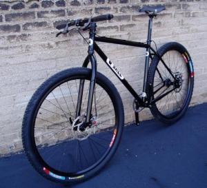 QBall Bikes QBall: A black mountain bike with wide tires, featuring a sleek frame and a minimalist design, leaning against a textured light-colored brick wall.