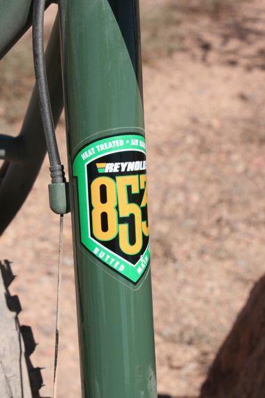 Raleigh XXIX Pro: Close-up image of a green bicycle frame featuring a Reynolds 853 sticker. The sticker highlights specifications like "Heat Treated" and "Air Hardened," emphasizing the quality and durability of the frame material. The background includes a blurred, natural terrain, suggesting outdoor use.