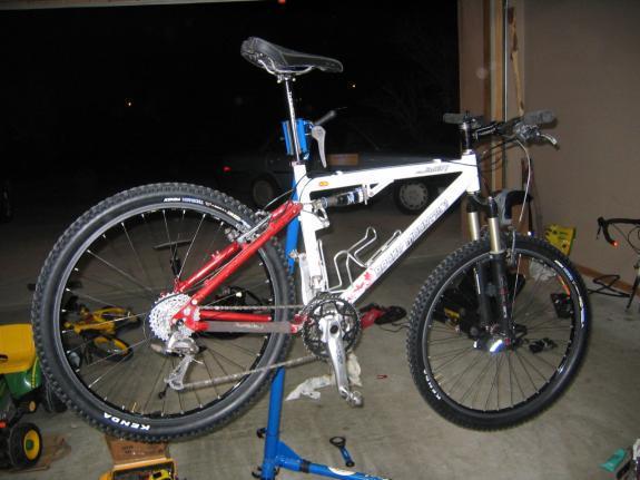 Rocky Mountain Element Team: A mountain bike in a workshop, mounted on a bike repair stand. The bike features a white frame with red and black accents, and has large tires. Various tools and equipment are scattered around the garage floor. The garage is dimly lit, indicating it is nighttime.