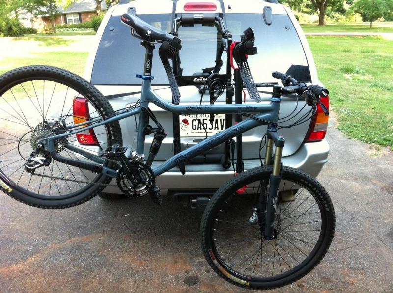 Salsa El Kaboing: A mountain bike secured on a bike rack attached to the back of an SUV. The bike has wide tires and a dual suspension frame, with the handlebars and front fork visible. The vehicle's rear is shown with a visible license plate and a grassy area in the background.