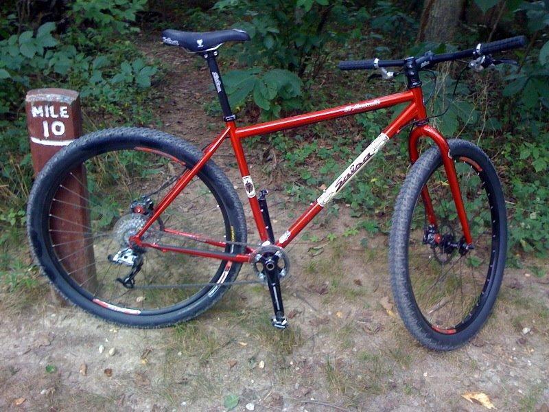 Salsa El Mariachi: A red mountain bike is positioned on a dirt trail next to a wooden mile marker sign that reads "MILE 10." The bike features thick tires suitable for off-road terrain and a visible gear system. Surrounding the bike is dense greenery, typical of a wooded area.
