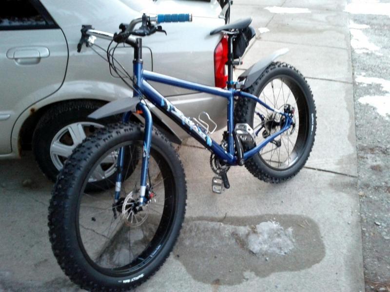 Salsa Mukluk: A blue fat bike with large tires parked alongside a car on a concrete surface. The bike features a sturdy frame, disc brakes, and a kickstand, with the handlebars equipped with blue grips. Snow and ice patches are visible on the ground.