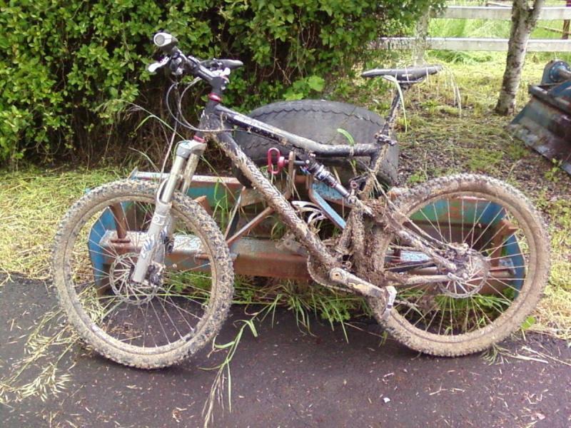 Santa Cruz Blur LT: A muddy mountain bike parked beside an old metal cart, surrounded by grass and bushes. The bike shows signs of recent use, with dirt and mud covering its frame and tires.