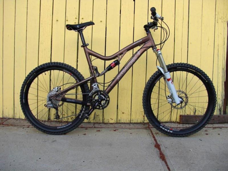 Santa Cruz Blur LT: A brown mountain bike with a silver front fork parked against a yellow wooden wall. The bike features thick tires, a dual suspension frame, and a gear system on the back wheel.