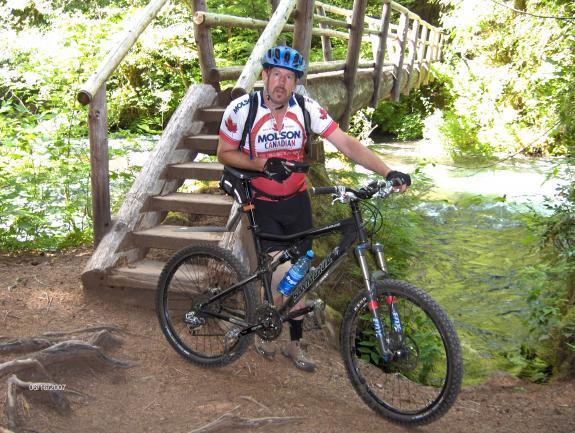 Santa Cruz Blur LT: A mountain biker wearing a helmet and cycling gear stands next to his bike beside a wooden bridge. The scene is surrounded by lush greenery and a flowing stream, suggesting a natural outdoor environment.