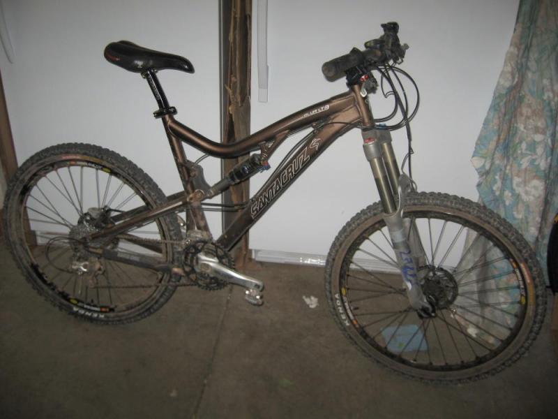 Santa Cruz Blur LT2: Mountain bike leaning against a wall, featuring a brown frame with the brand name "Santa Cruz." The bike has wide tires with visible tread, a front suspension fork, and a slightly dusty appearance, indicating recent outdoor use.