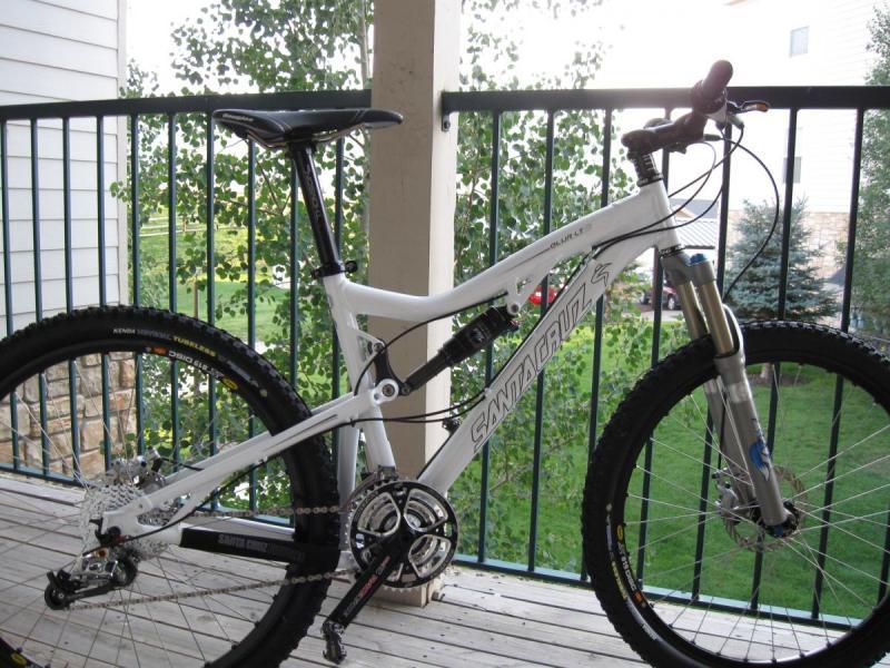 Santa Cruz Blur LT2: A white mountain bike with a sturdy frame, featuring front suspension and wide tires, is parked on a wooden balcony. The background shows greenery and a clear view of the outdoors.
